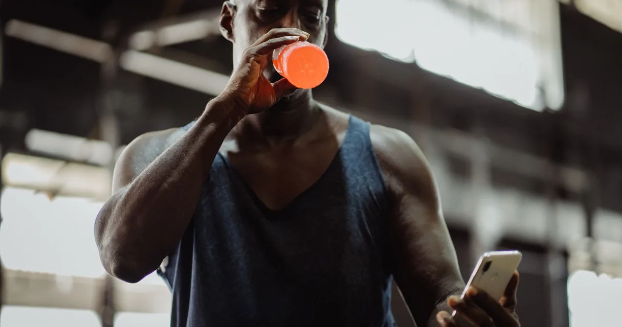 Homem bebendo água de uma garrafa laranja enquanto confere o celular na academia